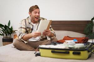 Portrait of man looking with excitement at his notebook, reading notes, going through check list of items, packing for holiday, sitting near opened suitcase photo