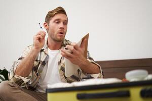 Portrait of tourist packing suitcase, going through check-list, writing down items to take with him in luggage, making notes in notebook, sitting on bed photo