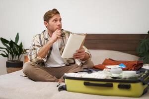 retrato de pensativo joven hombre, turista planificación su vacaciones, sentado con abierto maleta en cama, haciendo notas, escritura a hacer cheque lista en cuaderno foto