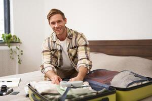 Portrait of young handsome bearded guy, tourist going on holiday, fastening his clothes in suitcase, packing for vacation, going on business trip, sitting in bedroom photo