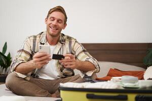 retrato de joven hombre, turista pago en línea, utilizando crédito tarjeta y móvil teléfono aplicación, sentado en cama con maleta, ordenando en Internet, comprando fiesta Entradas foto