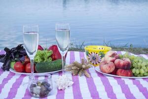 Beautiful picnic with fruits, vegetables and glasses of wine near lake. photo