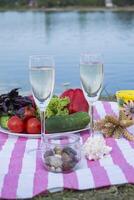 Beautiful picnic with fruits, vegetables and glasses of wine near lake. photo