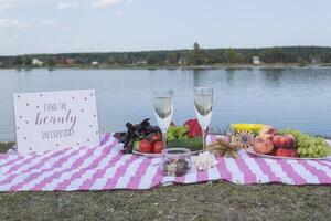 Beautiful picnic with fruits, vegetables and glasses of wine near lake. photo