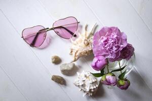 Pink glasses, peonies and sea shells on a white wooden table. Romantic summer flat lay. photo