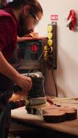 Vertical African american carpenter in joinery watching apprentice using orbital sander on lumber. Team in woodworking shop using angle grinder to create wooden decorations, refining wood objects, camera A video