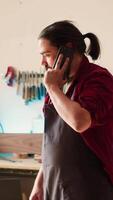 Vertical Cabinetmaker receiving telephone call from client inquiring about furniture assembling commission progress. Carpenter in assembly shop talking on smartphone with customer, camera A video