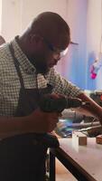 Vertical African american man in carpentry shop using power drill to create holes for dowels in wooden board. Craftsperson sinks screws into wood block with electric tool, doing precise drilling, camera B video