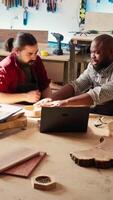 Vertical Carpenter and colleague verifying finished wood art product, comparing with blueprint. Artisan and apprentice inspecting wooden object for damages based on schematics on laptop screen, camera B video