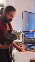 Vertical African american carpenter in joinery watching apprentice using orbital sander on lumber. Team in woodworking shop using angle grinder to create wooden decorations, refining wood objects, camera B video