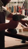 Vertical African american man in carpentry shop using power drill to create holes for dowels in wooden board. Craftsperson sinks screws into wood block with electric tool, doing precise drilling, camera A video