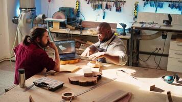 Carpenter looking over blueprints schematics on laptop in CAD software with apprentice. Woodworkers designing custom 3D furniture on notebook in studio, preparing for client order, camera B video