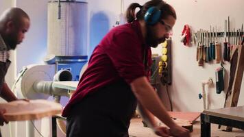 Craftsperson at work consulting blueprint and using orbital sander with sandpaper on lumber. Woodworking expert in carpentry shop uses angle grinder on wood after checking schematics, camera A video