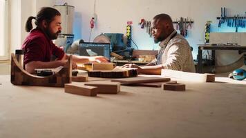 Artisan and apprentice comparing wood art piece with schematic displayed on CAD software on laptop. Woodworking experts in studio crosschecking wooden object with notebook blueprint, camera B video