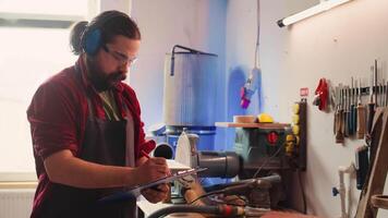 Craftsperson at work consulting blueprint and using orbital sander with sandpaper on lumber. Woodworking expert in carpentry shop uses angle grinder on wood after checking schematics, camera B video