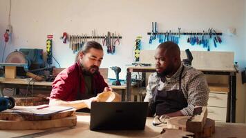 Woodworker looking over blueprints on laptop, brainstorming with african american apprentice. Team of manufacturers discussing furniture assembly schematics on notebook in studio, camera B video