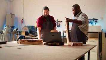 Woodworker looking over blueprints on laptop, brainstorming with colleague next steps in wood processing. Manufacturer and BIPOC apprentice reading furniture assembly schematics in studio, camera B video