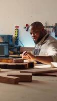 Vertical Happy BIPOC cabinetmaker and colleague comparing wood piece with schematic displayed on laptop. Cheerful carpenters in joinery crosschecking wooden object with notebook blueprint video