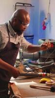 verticaal beeldhouwer vormgeven rauw hout gebruik makend van beitel en hamer in timmerwerk winkel met collega schoonmaak werkbank in achtergrond. bipoc Mens maken hout beeldhouwwerken, gravure plank met hulpmiddelen, camera b video