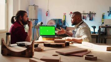Woodworker looking over blueprints on green screen laptop in CAD software with apprentice. Craftsperson and coworker designing custom 3D furniture on chroma key notebook in studio, camera B video
