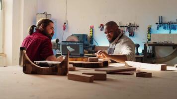 Happy BIPOC cabinetmaker and colleague comparing wood piece with schematic displayed on laptop. Cheerful carpenters in joinery crosschecking wooden object with notebook blueprint video