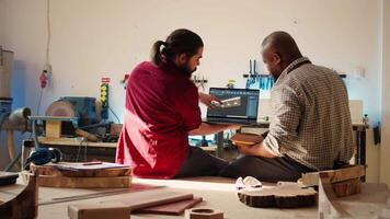 African american manufacturer and colleague comparing wood art piece with schematic displayed on CAD software on laptop. Woodworking professionals crosschecking wooden object with blueprint, camera B video