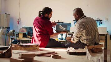 Woodworker looking over blueprints on laptop in CAD software with apprentice. Manufacturer and coworker designing custom 3D furniture on notebook in studio, preparing for customer order, camera B video