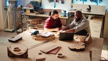 snickare och kollega verifierande färdiga trä konst produkt, jämförande med plan. hantverkare och lärling inspekterande trä- objekt för skadestånd baserad på scheman på bärbar dator skärm, kamera b video