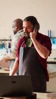 Vertical Cabinetmaker receiving telephone call from client inquiring about furniture assembling commission progress. Carpenter in assembly shop talking on smartphone with customer, camera B video