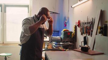 African american man in woodworking shop inspecting lumber piece before assembling furniture, checking for scratches. Cabinetmaker evaluates timber block, ensuring it meets quality standards, camera B video