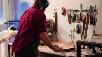 Carpenter at work consulting blueprint, using orbital sander on lumber, wearing safety glasses. Artisan with protecting equipment uses angle grinder on wood after checking schematics, camera A video