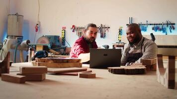 Cabinetmakers doing brainstorming while using CAD software on laptop, discussing wood designs. Carpenters in assembly shop discussing, using program on notebook to plan furniture assembling, camera B video