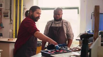 Master in carpentry shop instructing BIPOC worker to wear protection gear before assembling furniture. Cabinetmaker making sure apprentice uses safety earmuffs and glasses before starting work, camera B video