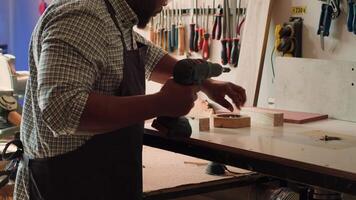 African american man in carpentry shop using power drill to create holes for dowels in wooden board. Craftsperson sinks screws into wood block with electric tool, doing precise drilling, camera A video