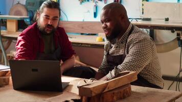 Cabinetmakers doing brainstorming while using CAD software on laptop, discussing wood designs. Carpenters in assembly shop discussing, using program on notebook to plan furniture assembling, camera A video