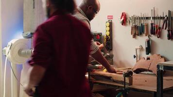 Sculptor shaping raw timber using chisel and hammer in carpentry shop with coworker cleaning workbench in background. BIPOC man making wood sculptures, engraving plank with tools, camera A video