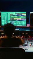 Vertical African american sound designer editing audio recordings in control room, working with switchers and faders on mixer console. Music producer creating new tunes and tracks. Camera B. video