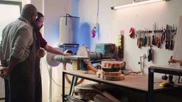 Manufacturer teaching new employee how to shape wooden bowl on disc sander in assembling shop. Apprentice learning from woodworking specialist how to use grinding machine rotating disc, camera B video