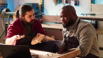 Woodworker looking over blueprints on laptop, brainstorming with african american apprentice. Team of manufacturers discussing furniture assembly schematics on notebook in studio, camera A video