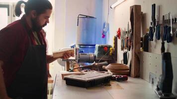 Cabinetmaker drills hole into lumber block, doing furniture assembling in studio, close up. Manufacturer in assembly shop inserting nail into piece of wood using electric power drill, camera B video