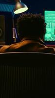 Vertical African american audio engineer using sound mixing console for managing tracks, music post production. Producer pressing motorized control knobs, looks at editing software. Camera B. video