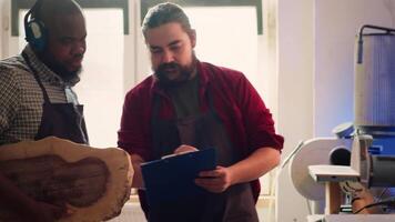 carpintero mirando terminado planos en computadora portátil, lluvia de ideas con colega siguiente pasos en madera Procesando. fabricante y bipoc aprendiz leyendo mueble montaje esquemas en estudio, cámara un video