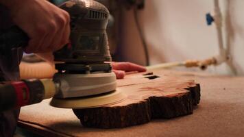 Craftsperson works with orbital sander on lumber block to achieve smooth finish, close up shot. Woodworking specialist in furniture assembly shop using angle grinder to fix wooden surface, camera A video