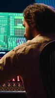 Vertical Sound designer mixing and mastering tracks on audio console in control room, using daw software on computer and editing files. Technician creating new music in professional studio. Camera A. video