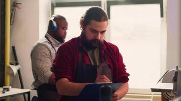 Craftsperson drawing blueprints on notepad in studio with BIPOC coworker in background solving tasks. Manufacturer looking at technical schematics to execute woodworking projects, camera A video