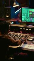 Vertical African american audio expert adding sound effects on tracks in post production, operating mixing console and pushing sliders. Producer creating music for his new album, audio mixer. Camera B. video