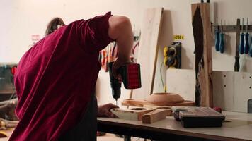 Woodworker in assembly shop using power drill to create holes for dowels in wooden board. Carpenter sinking screws into wooden surfaces with electric tool, doing precise drilling for seamless joinery, camera A video