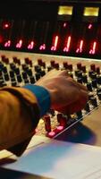 Vertical Audio technician producing new music at mixing console in control room, twisting knobs, pressing sliders and switchers. Producer working on processing and editing sounds quality. Camera A. video