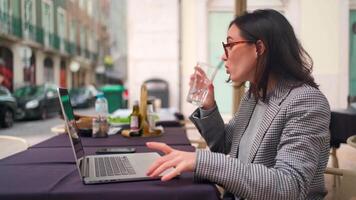 a woman in glasses drinking water from a glass while sitting at a table with a laptop video