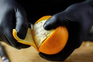 Person with black gloves peeling full orange on wooden cutting board photo